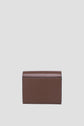 A small, rectangular brown leather wallet from Gerard Darel is displayed standing upright against a plain light gray background. This portefeuille cuir compact is closed, showcasing its smooth exterior with visible stitching along the edges.