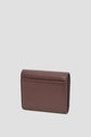 A brown Gerard Darel portefeuille cuir is standing upright on a plain, light gray background. The wallet is closed, showing its smooth back and simple, minimalist design.