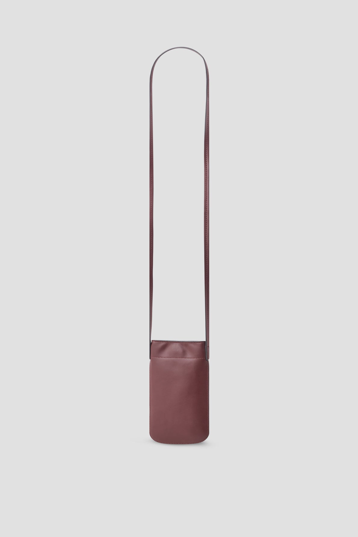 A minimalist, burgundy leather crossbody bag with a long, thin strap—perfect as a porte-téléphone cuir—is displayed against a plain light gray background.