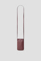A minimalist, burgundy leather crossbody bag with a long, thin strap—perfect as a porte-téléphone cuir—is displayed against a plain light gray background.
