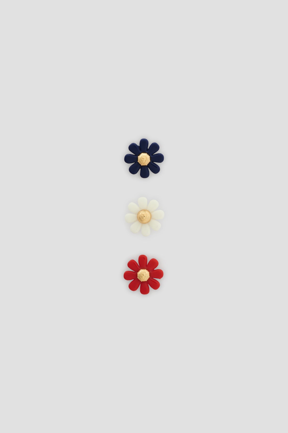 Three daisy-shaped objets, each with a yellow center, are arranged vertically on a light gray background. Reminiscent of fleurs bijoux from the Bijoux Darel collection, the top flower is navy blue, the middle is white, and the bottom is red—a charming accessory that can add an elegant touch to chaussures or outfits alike.