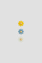 Three flower-shaped objects are arranged vertically on a light gray background: yellow at the top, blue in the middle, and pale yellow at the bottom. Each flower has a beige circular center, reminiscent of délicate fleurs bijoux often seen in bijoux Darel collections.