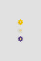Three daisy-shaped objets reminiscent of fleurs bijoux, with wooden centers arranged vertically: yellow on top, white in the middle, and purple at the bottom, against a plain light gray background.