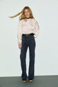 A woman with long blonde hair wears a pale satin blouse, dark monogramme taille haute jeans flare, a black belt, and platform shoes, standing against a plain light background.
