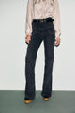 A person wearing a light long-sleeve blouse, high-waisted dark patterned pants taille haute, a black belt, and platform shoes stands against a light-colored wall. Their head is not visible in the image.