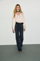 A woman with long blonde hair wears a light pink blouse, dark jean flare monogramme taille haute, a black belt, platform shoes, and a black choker, walking against a plain white background.