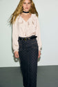 A person with long wavy blonde hair wears a cream ruffled blouse, a black patterned taille haute skirt cinched with a black belt, and a black choker, standing against a plain light background.