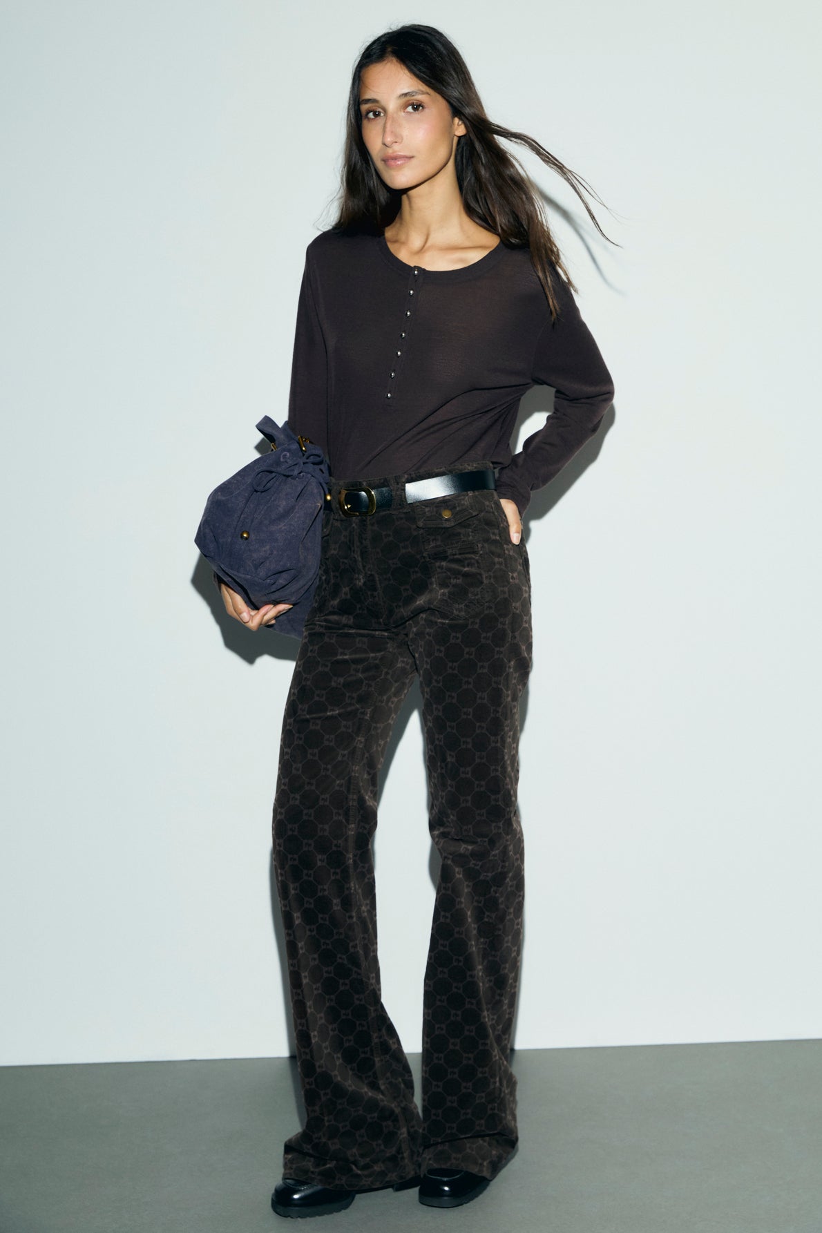 Woman standing against a light-colored wall, wearing a dark long-sleeve shirt, patterned pantalon velours marron, black belt with monogramme GD detailing, and holding a dark bag; her hair is loose and she is looking slightly to the side.