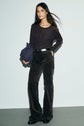 Woman standing against a light-colored wall, wearing a dark long-sleeve shirt, patterned pantalon velours marron, black belt with monogramme GD detailing, and holding a dark bag; her hair is loose and she is looking slightly to the side.