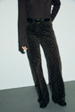 A person wearing a dark long-sleeve top and high-waisted, patterned pantalon velours marron with a belt stands against a light background; only their torso and legs are visible.