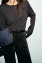 Woman in a dark long-sleeve top and patterned high-waisted pants with a black belt, holding a blue suede handbag, standing against a plain light background. Her head is partially out of frame. The look channels effortless style with hints of retro inspiration, perfect for pairing with pantalon velours marron or classic jean flare femme silhouettes.