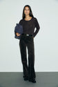 A woman stands against a plain wall, wearing a black long-sleeve top, a pantalon velours marron with a flared cut, black belt, and holding a blue bag under her arm.