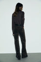Woman standing with her back to the camera, wearing a dark long-sleeve top and high-waisted, patterned jean flare femme. The background is plain and light-colored.