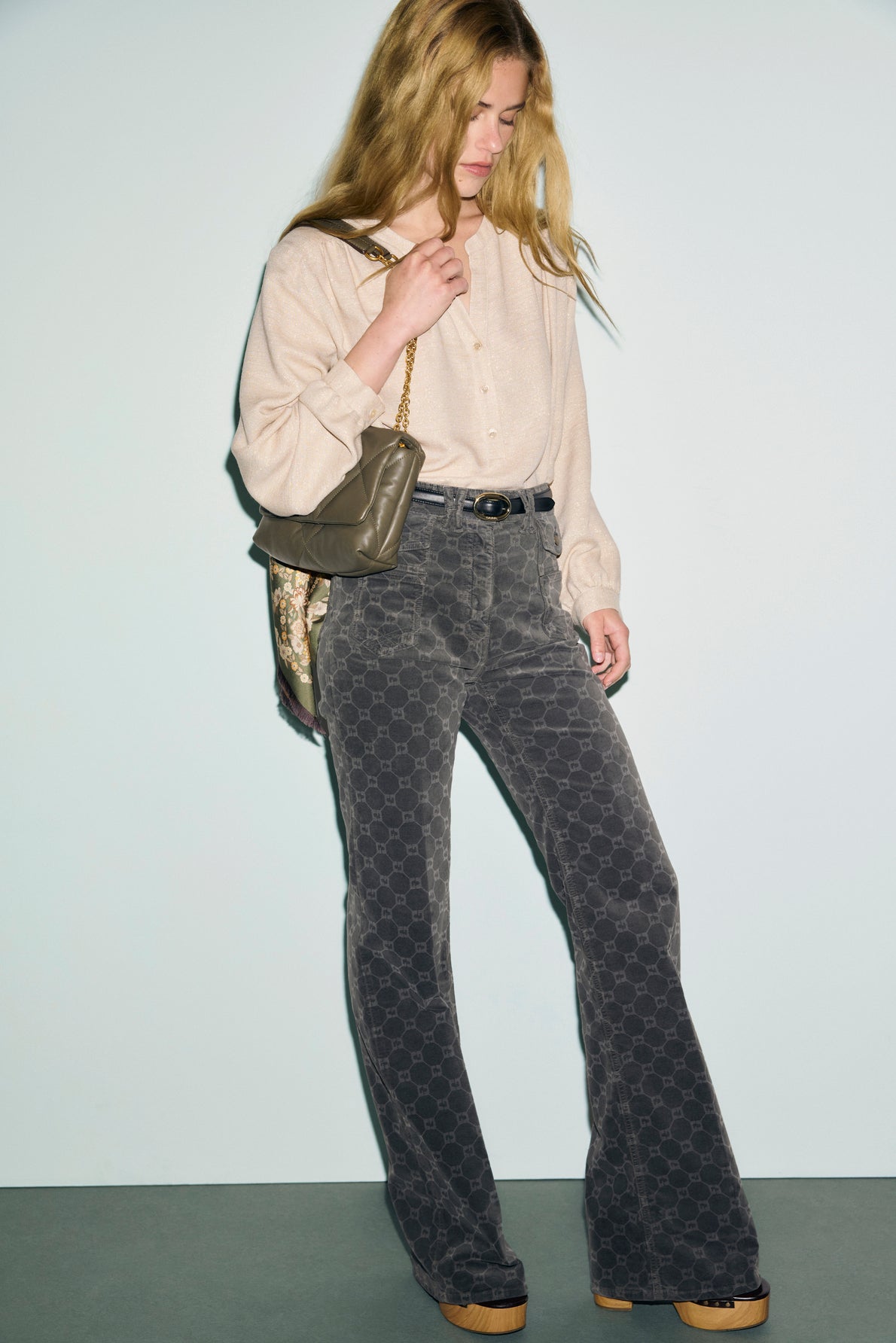 A woman with long blonde hair wears a beige blouse, a pantalon taille haute with a striking pattern, platform shoes, and carries a quilted shoulder bag while looking down.
