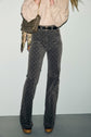 Person wearing a pantalon taille haute in patterned gray, a light beige shirt, and holding a taupe handbag with a floral scarf. Face is not visible. The high-waisted pants have a subtle geometric print reminiscent of motif monogramme details, and the shoes are platform style.