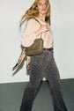 A woman with long blonde hair walks, wearing a beige blouse, patterned gray pantalon taille haute, a black belt, and carrying a quilted olive green shoulder bag with a floral scarf tied to it.