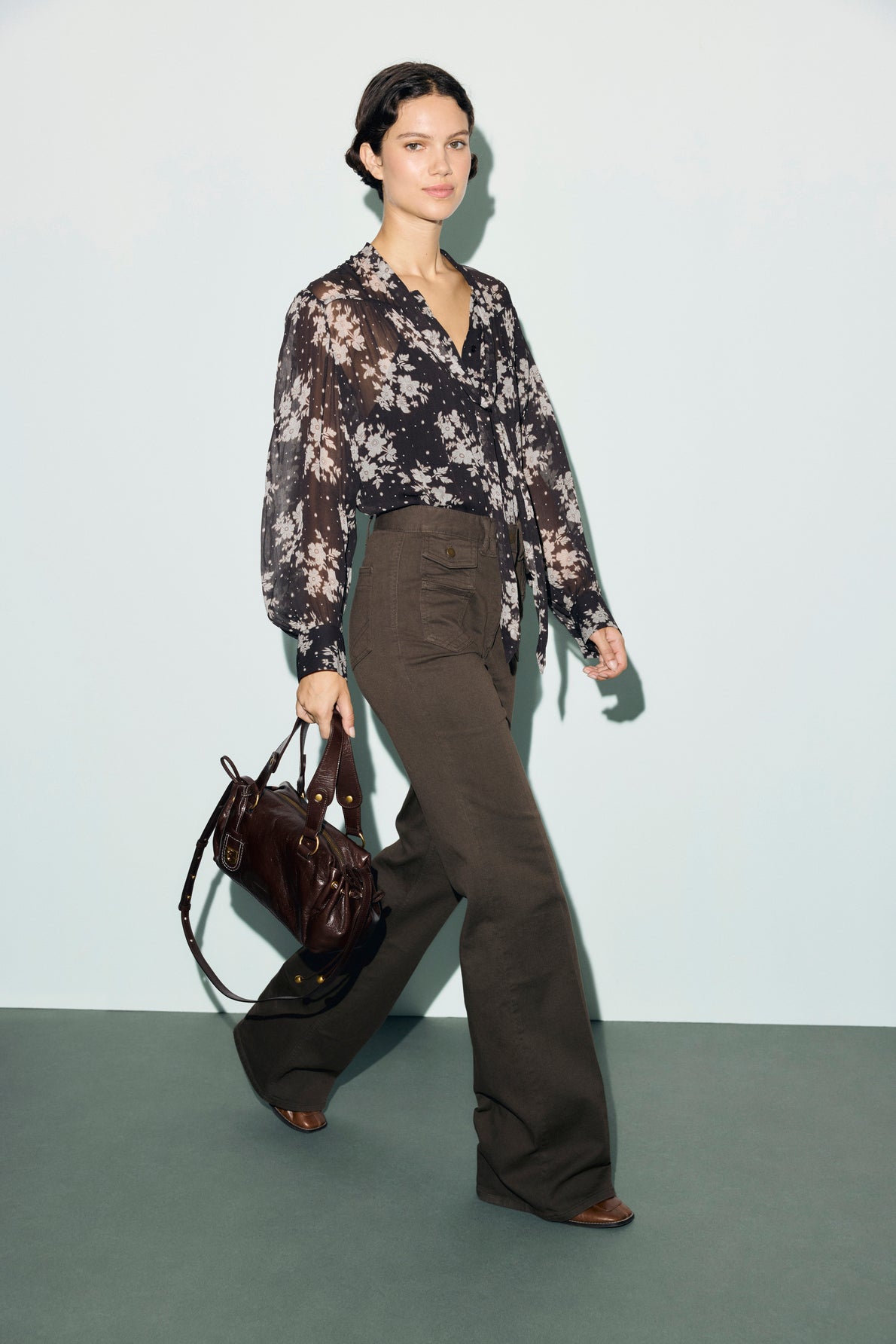 A woman wearing a sheer floral blouse and wide-leg, taille haute pantalon marron walks forward, holding a dark brown handbag. She has short dark hair and wears brown shoes. The background is plain and minimalist.