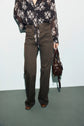 Person wearing a dark floral blouse and high-waisted olive green trousers, styled in a flattering taille haute cut, holding a brown handbag by their side, standing against a light background. Only the lower face, torso, and legs are visible.