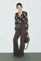 A person stands against a plain background wearing a black floral blouse, a pantalon marron taille haute with a wide-leg jean flare silhouette, and holding a brown handbag.