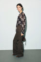 A person stands against a light background wearing a sheer black floral blouse and high-waisted wide-leg brown pantalon marron, holding a small brown handbag.