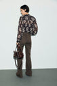 A person stands facing away, wearing a sheer black floral blouse and a pantalon marron taille haute, holding a dark brown handbag in their left hand against a plain background.