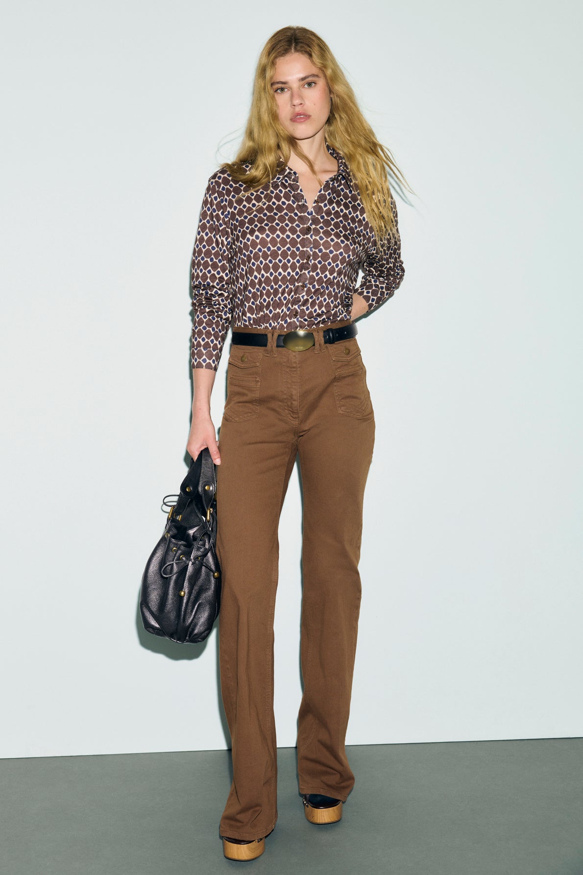 A woman with long blonde hair wears a patterned shirt, pantalon cannelle taille haute, platform shoes, and holds a black handbag, standing against a plain light background.