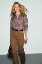 A woman with long, wavy blonde hair wears a patterned brown blouse, a pantalon cannelle with a black belt, and brown pants. She carries a black bag and walks confidently against a light background.