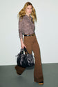 A woman with long blonde hair wears a patterned shirt, pantalon cannelle flare taille haute, and brown shoes, carrying a large black handbag while walking against a light background.