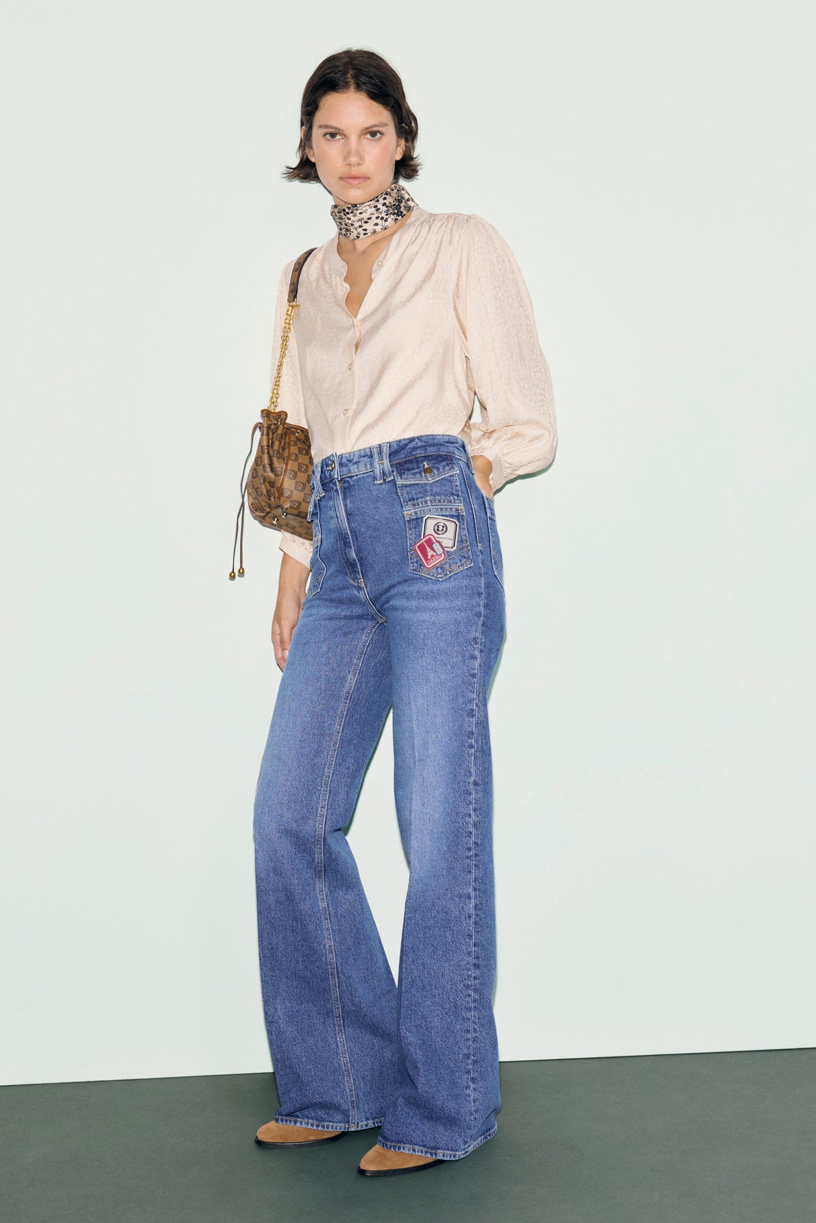 Woman wearing a cream blouse, blue jean flare taille haute with patches, tan shoes, a scarf around her neck, and a brown shoulder bag, standing against a plain light background.