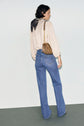 A person with short dark hair, wearing a cream blouse, blue jean flare taille haute, a black scarf, and carrying a small brown shoulder bag, stands facing away from the camera against a plain background.