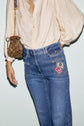 A person wearing high-waisted blue jeans with patches and a cream blouse, carrying a small brown handbag with a gold chain strap. The jean flare silhouette and pantalon long style add a retro-chic touch to the overall look.