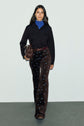 A woman with long dark hair wears a blue turtleneck under a black shirt, paired with taille haute pantalon flare velours à motif fleuri and brown shoes. She carries a brown handbag and walks on a gray floor against a plain light background.
