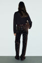 A person with long brown hair stands facing away, wearing a black top and high-waisted pantalon flare velours with a motif fleuri. They hold a brown handbag and wear a dark belt. The background is plain and light-colored.