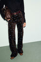 A person wearing a black top, a taille haute pantalon flare velours in brown with blue motif fleuri, a maroon belt, brown shoes, and holding a brown leather bag, stands against a pale background.