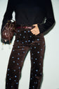 Person wearing a black long-sleeve top and a pantalon flare velours taille haute in brown, detailed with small blue motif fleuri, holding a brown handbag. Only the torso and legs are visible.