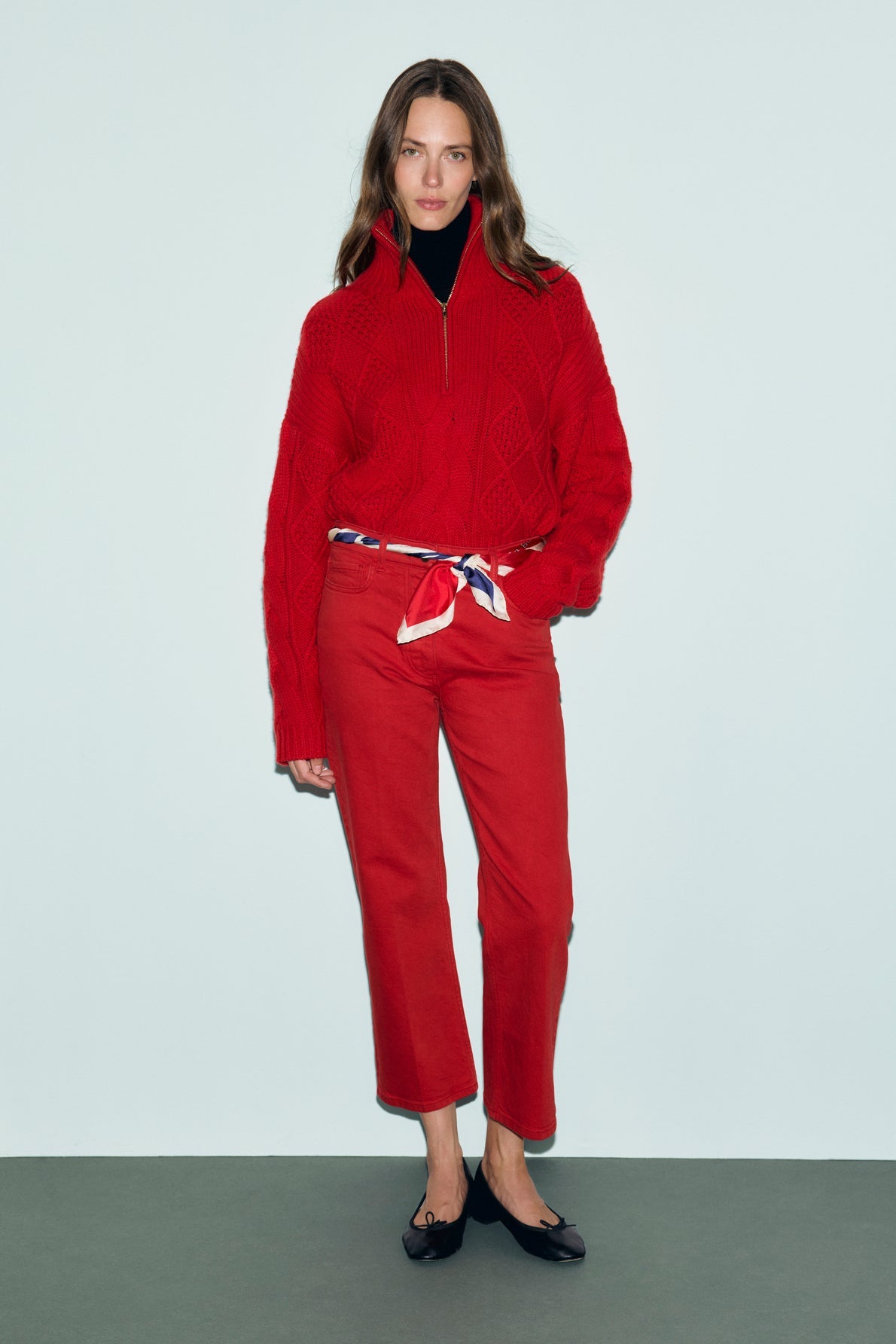 A woman stands against a plain background wearing a red coupe droite jean 7/8, a matching red sweater, a patterned scarf belt, black flats, and a black turtleneck underneath.