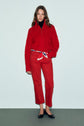 A woman stands against a plain background wearing a red coupe droite jean 7/8, a matching red sweater, a patterned scarf belt, black flats, and a black turtleneck underneath.