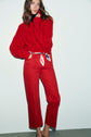 A person stands against a white wall wearing a vibrant red cable knit sweater paired with a jean rouge coupe droite and a chic scarf belt. They have long brown hair, and their left hand touches their chin while they look at the camera.