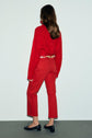 A woman with long brown hair faces away, wearing a textured red sweater, jean rouge coupe droite 7/8, a printed belt, and black low-heeled shoes. She stands against a plain, light background.