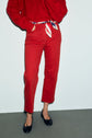 Person wearing a red sweater and coupe droite jean rouge with a striped scarf belt, standing on a gray floor. Only the lower half of the body is visible. The person is also wearing black ballet flats.