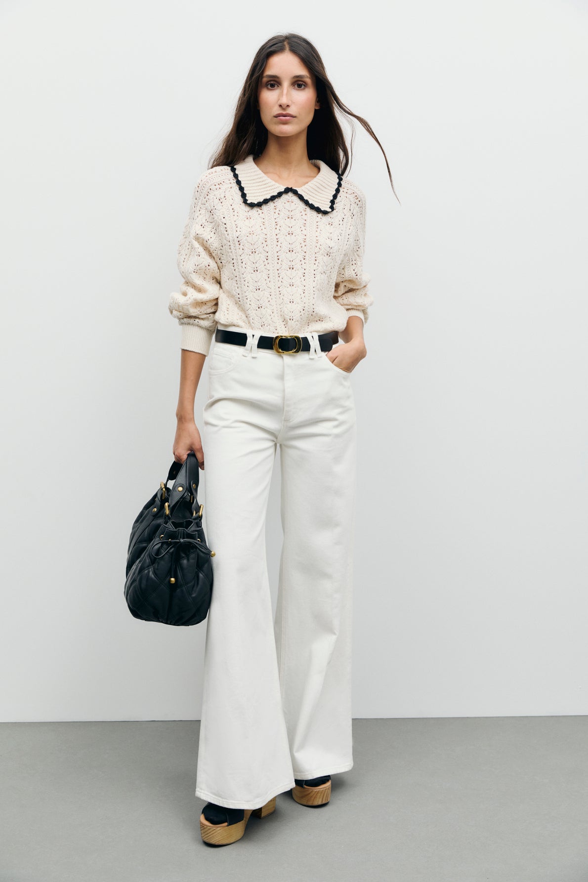 A woman stands against a plain wall wearing a cream knit sweater with a black-trimmed collar, coupe droite wide-leg white pants from the collection Studio, platform shoes, and holding a black handbag.