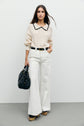 A woman stands against a plain wall wearing a cream knit sweater with a black-trimmed collar, coupe droite wide-leg white pants from the collection Studio, platform shoes, and holding a black handbag.