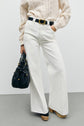 Person wearing a cream knit sweater, high-waisted jean blanc coupe droite from the collection Studio, paired with a black belt, wooden platform shoes, and holding a black handbag with gold details, standing against a plain light background.