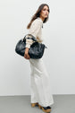 Woman in a cream knit sweater and jean blanc flared pants holds a large black quilted bag. She wears platform heels and stands against a plain light gray background, effortlessly channeling the modern aesthetic of the collection Studio with her chic coupe droite silhouette.