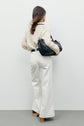 A woman stands facing away, wearing a cream knit sweater, a pair of jean blanc coupe droite from the collection Studio, tan heeled shoes, and holding a large black handbag over her shoulder against a plain white background.