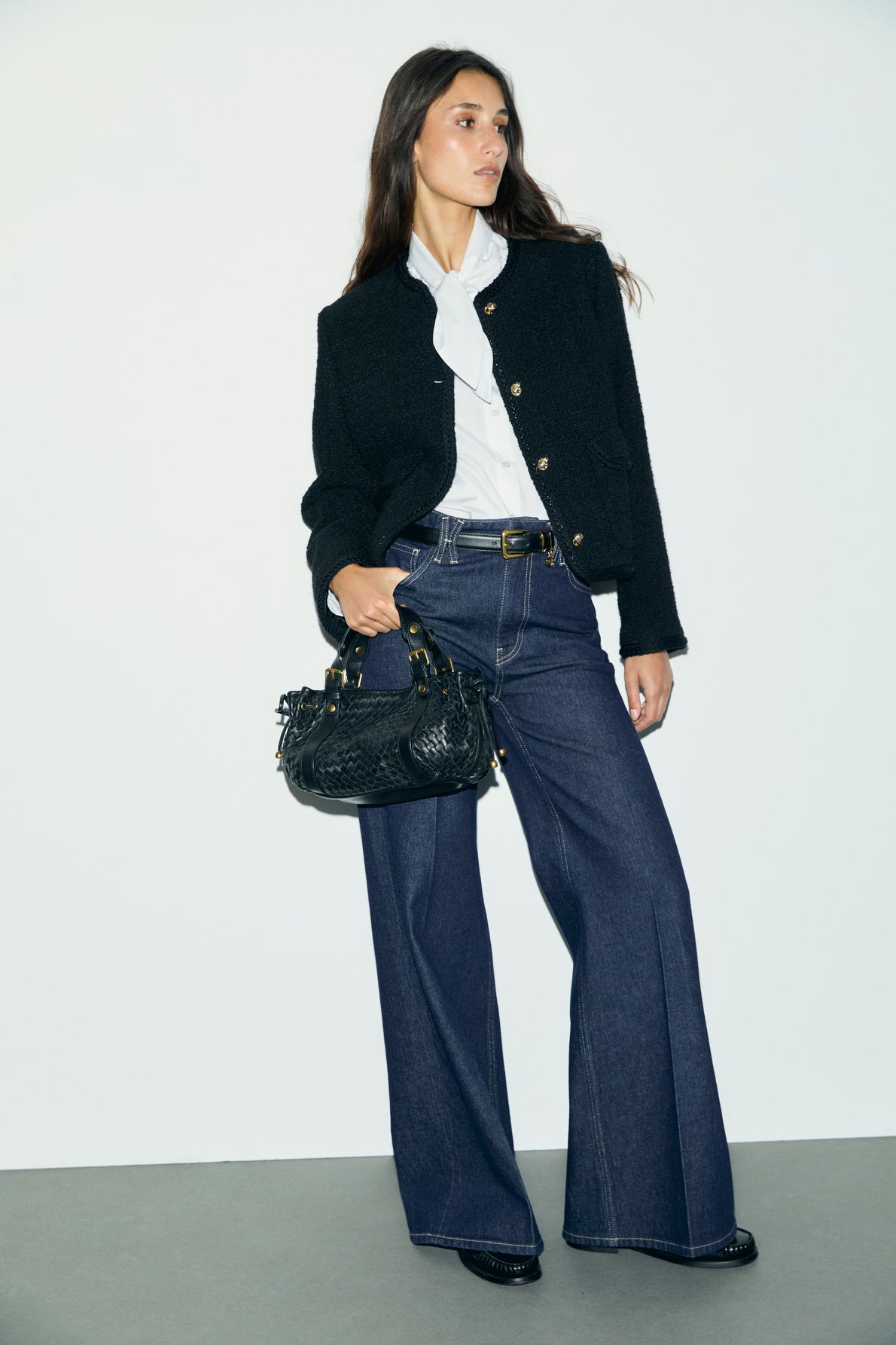 A woman in a black jacket, white blouse, and wide-leg bleu jean brut stands holding a black handbag. She has long brown hair and is posing against a plain white wall and gray floor, showcasing the modern allure of une coupe large.