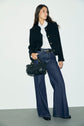 A woman in a black jacket, white blouse, and wide-leg bleu jean brut stands holding a black handbag. She has long brown hair and is posing against a plain white wall and gray floor, showcasing the modern allure of une coupe large.