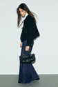 Woman with long brown hair wears a black jacket, white blouse, and wide-leg coupe large jeans. She holds a black woven handbag and looks down while standing against a plain white wall.