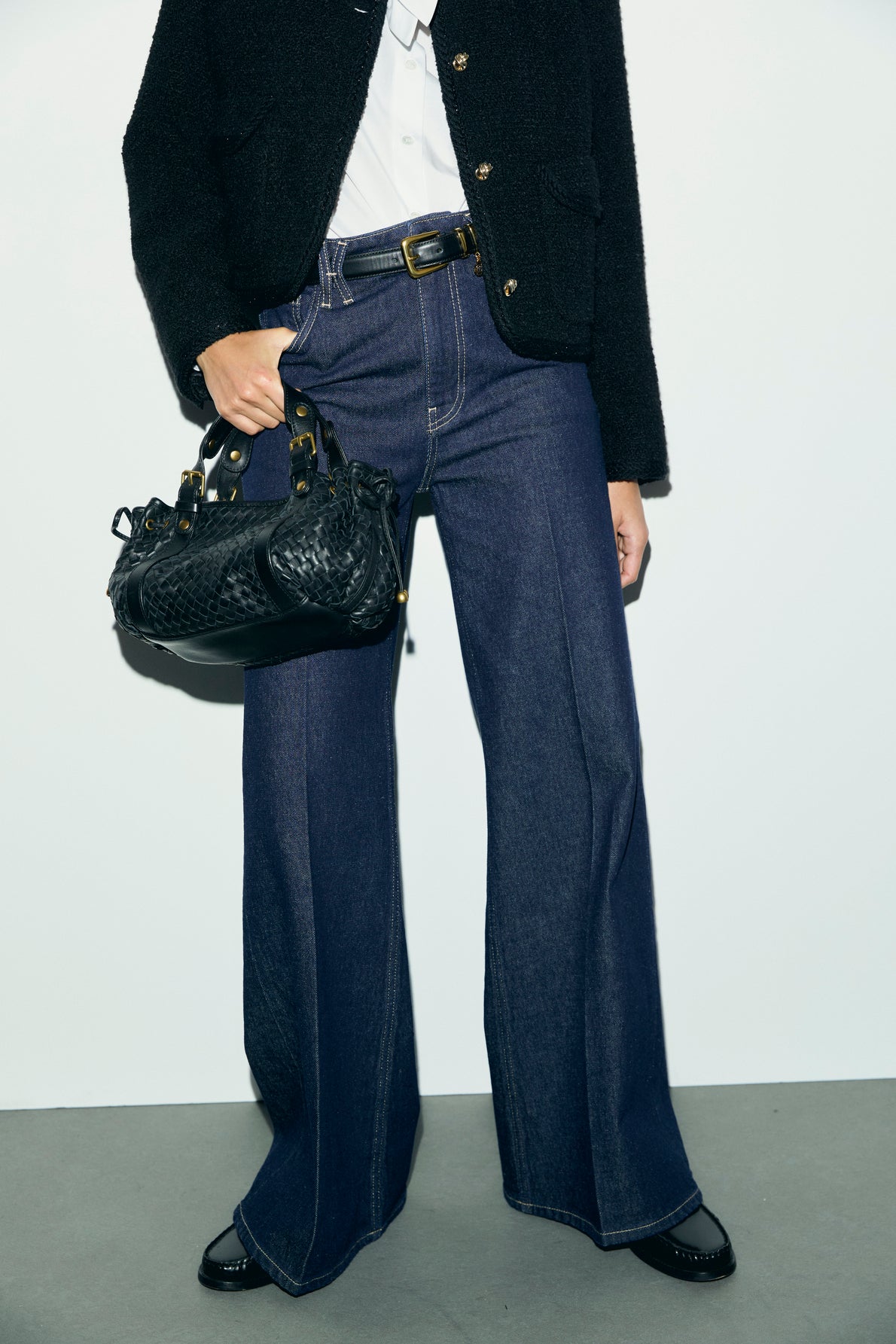 A person wearing a textured black jacket, white shirt, wide-leg dark blue jeans à coupe large, and black shoes holds a black handbag with gold hardware, standing against a plain light background.