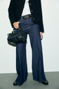 A person wearing a textured black jacket, white shirt, wide-leg dark blue jeans à coupe large, and black shoes holds a black handbag with gold hardware, standing against a plain light background.
